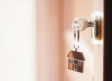 A key in the lock of a slightly open apartment door, with a house-shaped keychain hanging from the key