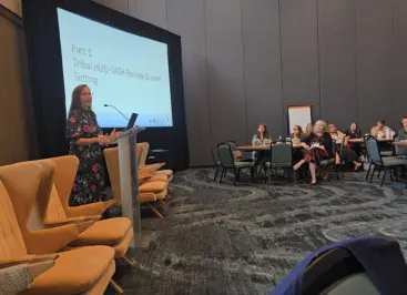 A woman in a flowered dress speaking to a conference audience; she is standing in front of a slide that says 