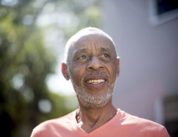 Elderly man in a pink t-shirt, smiling outside an apartment building on a sunny day