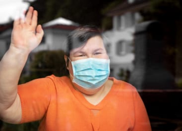 Friendly woman in orange shirt wearing a surgical mask and waving through a closed window