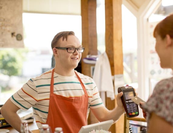 In a sunny cafe, a young adult barista with an intellectual disability accepts payment for a purchase from a customer