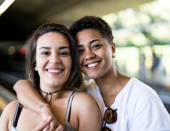 Two smiling young women in an affectionate embrace on an outdoor train platform