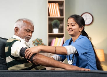 A smiling doctor or nurse in scrubs, taking an elderly man's blood pressure in his home.
