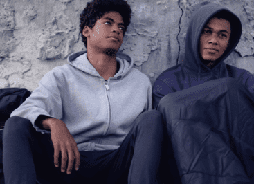 Two African American teen boys sit together on the ground, leaning against a rough concrete wall. Both wear hoodies; one has his hood up and a blanket over his knees.