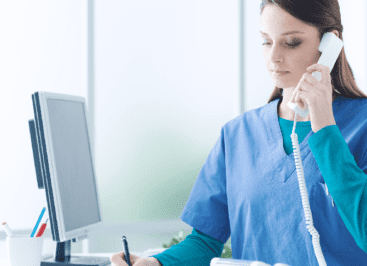 A nurse in scrubs is taking notes while listening to someone on the phone