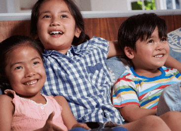 Three smiling siblings sit comfortably on a couch; 8-year-old her arms loosely over the shoulders of the younger ones on either side, who are 3 and 4 years old