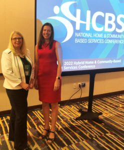 Nicole LiBaire and Lori Gerhard standing together in front of a large screen showing the HCBS conference logo