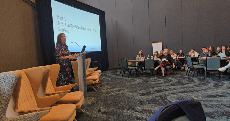 A woman in a flowered dress speaking to a conference audience; she is standing in front of a slide that says "Tribal HUD-VASH Review & Level Setting"