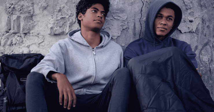 Two African American teen boys sit together on the ground, leaning against a rough concrete wall. Both wear hoodies; one has his hood up and a blanket over his knees.